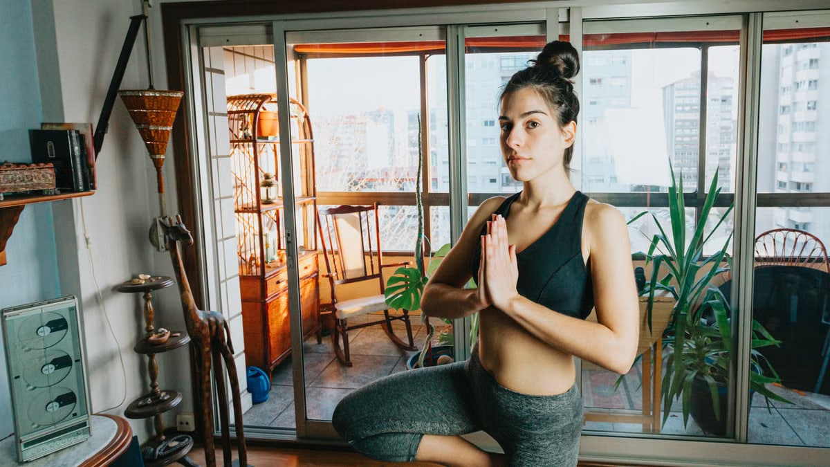 A young woman practices yoga indoors, standing in tree pose by a large window with a city view.